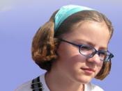 English: A young girl with glasses, dressed in a white shirt with a blue headband; blue sky in the background.