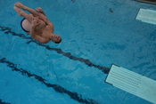 English: A male diver performs a reverse dive from the 3 meter springboard in the tuck position