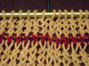 Knitted fabric showing a single red course of plaited stitches. The five stitches to the right of the blue needle at the top were knit into the back loop to twist the stitches one way; the five to the left of the same needle were knit by wrapping the yarn
