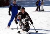 A person without the use of his legs learning to ski on a sit-ski, using two outriggers.