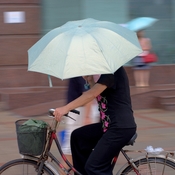 English: A woman is riding her bike under the rain and hold an umbrella with her hand. Nanjing, China.