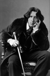English: Oscar Wilde, three-quarter length portrait, facing front, seated, leaning forward, left elbow resting on knee, hand to chin, holding walking stick in right hand, wearing coat.